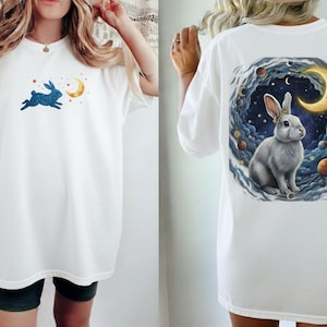 以下が含まれることがあります： 青いウサギが三日月と星に向かって跳ねる白いTシャツ。Tシャツの背面には、三日月と惑星のある天体の背景に、王冠をかぶった白いウサギが描かれています。