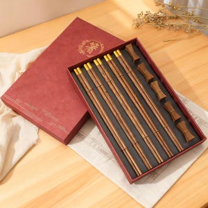 Könnte beinhalten: Ein Set aus Holz-Essstäbchen mit goldenen Akzenten, präsentiert in einer roten Geschenkbox. Die Essstäbchen sind in einem schwarzen Tablett angeordnet, zusammen mit passenden Essstäbchenhaltern. Die Box hat goldene Dekorationselemente.