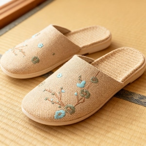 Può includere: Un paio di pantofole in lino beige con ricami floreali nei toni del blu e del verde. Le pantofole hanno un design semplice a punta aperta e una palette di colori neutri, adatte per l'uso in casa.