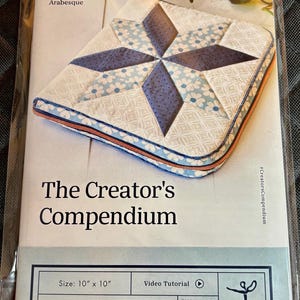 May include: A package featuring a quilted fabric project with a starburst design in shades of blue and white. The text "The Creator's Compendium" is displayed, along with size details (10" x 10") and pattern information. The difficulty rating is "Intrepid Adventure".