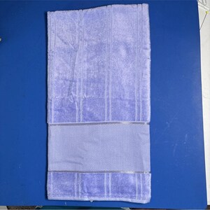 Pu&ograve; includere: Un asciugamano da bagno piegato, di colore viola chiaro, con una superficie strutturata e una fascia decorativa. L'asciugamano &egrave; rettangolare e presenta strisce verticali. Lo sfondo &egrave; una superficie blu.
