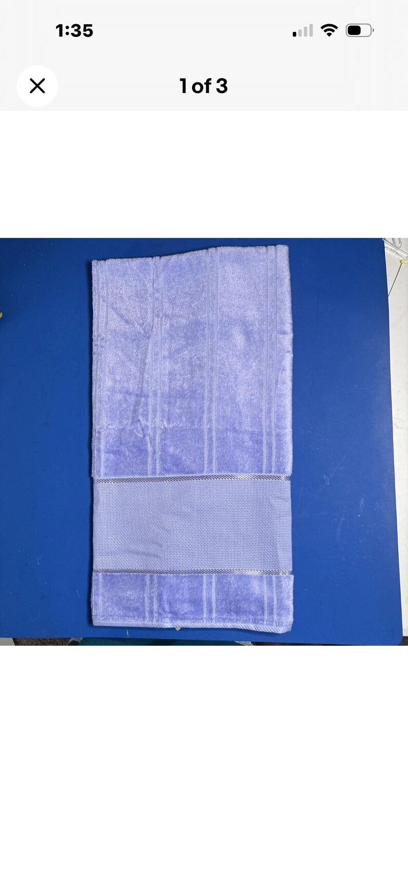 Pu&ograve; includere: Un asciugamano da bagno piegato, di colore viola chiaro, con una superficie strutturata e una fascia decorativa. L'asciugamano &egrave; rettangolare e presenta strisce verticali. Lo sfondo &egrave; una superficie blu.