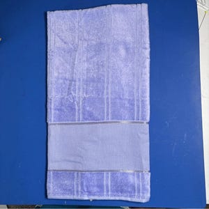 Pu&ograve; includere: Un asciugamano da bagno color lavanda con una superficie strutturata e una fascia decorativa. L'asciugamano &egrave; piegato e appoggiato su una superficie blu. L'asciugamano &egrave; di colore viola chiaro.