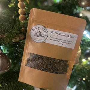 May include: A brown paper pouch of "Signature Blend" tea, with a clear window showing the tea leaves. The label includes the text "Pot & Petal" and a list of ingredients: Peppermint, Chamomile, Lavender, Lemon Balm, Calandula, Nettle, and Licorice. The pouch is displayed on a Christmas tree.