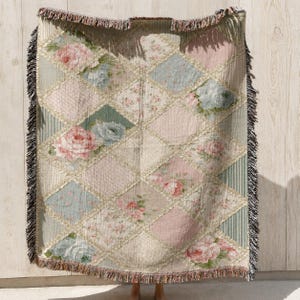 May include: A woven throw blanket featuring a diamond-patterned patchwork design with alternating squares of floral prints in shades of pink, blue, and cream, accented by vertical stripes and finished with a decorative fringed edge on all sides.