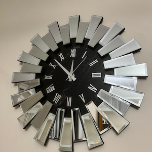 May include: A circular wall clock featuring a black face with silver Roman numerals. It has a sunburst design created by rectangular mirrored accents. The clock hands are silver.