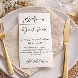 May include: A formal place setting with a white plate, gold fork and knife, and a linen napkin. A card with calligraphy in multiple languages is centered on the plate. The setting is decorated with flowers and a small bowl of olives.