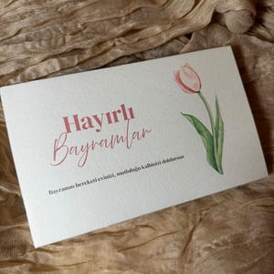 Può includere: Un biglietto di auguri bianco con la scritta "Hayırlı Bayramlar" in rosa, accompagnato da un'illustrazione di tulipano ad acquerello. Il biglietto è posto su uno sfondo di tessuto beige strutturato.
