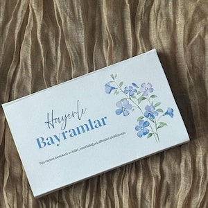 Pode incluir: Um cartão retangular branco com as palavras "Hayırlı Bayramlar" em escrita azul e um desenho floral de flores azuis. O cartão está sobre um fundo de tecido bege texturizado.
