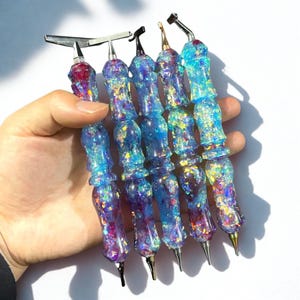 Könnte beinhalten: Eine Sammlung von Kunstwerkzeugen aus Harz mit schillerndem Glitzer. Die Werkzeuge haben ein blaues und violettes Farbschema mit silbernen Metallspitzen. Die Griffe sind mit einem strukturierten Design geformt. Die Werkzeuge sind für Bastel- und Kunstprojekte konzipiert.