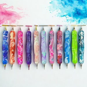 Op de afbeelding: Een verzameling van twaalf kleurrijke diamond painting pennen met gemarmerde hars handvatten. De pennen hebben verschillende kleuren, waaronder blauw, roze, paars en groen. Elke pen heeft een zilveren metalen punt.