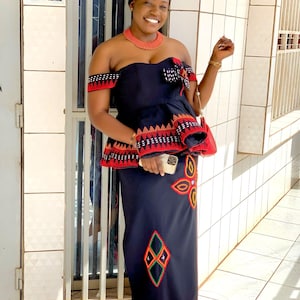 May include: A woman wearing a traditional black and red ensemble, including a strapless top and long skirt, adorned with intricate embroidery. She wears a matching hat and necklace, with a phone in her hand. The outfit features geometric and floral patterns.