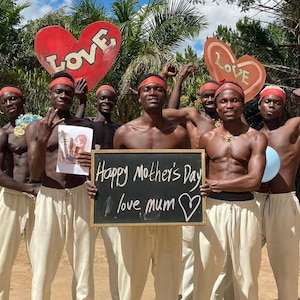 Könnte beinhalten: Eine Gruppe von Männern feiert den Muttertag mit Schildern und Requisiten. Sie tragen rote Stirnbänder und beige Hosen. Ein Schild zeigt "Happy Mother's Day love mum", ein anderes "LOVE" in Herzform.