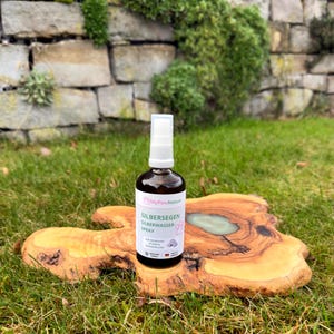 Könnte beinhalten: Eine braune Glassprühflasche mit einem weißen und rosa Etikett mit dem Text "Silbersegen Silberwasser Spray". Die Flasche steht auf einer Holzplatte mit hellgrüner Harzeinlage, vor dem Hintergrund von grünem Gras und einer Steinmauer.