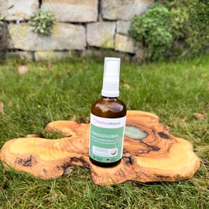 Könnte beinhalten: Eine braune Glassprühflasche mit weißem Deckel und einem grün-weißen Etikett mit der Aufschrift "MyPawNature KOKOSZAUBER". Die Flasche steht auf einer unregelmäßig geformten Holzoberfläche mit einem grün-braunen Hintergrund aus Gras und Stein.