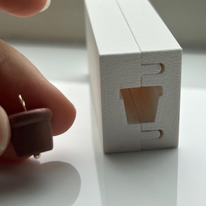 May include: A brown acorn-shaped earring with a gold-coloured post is held near a white, rectangular jewellery box. The box is made of two connected pieces with a cutout shaped like a small pot. The earring is a miniature representation of an acorn.