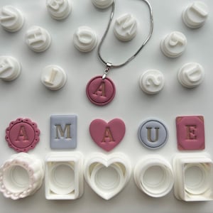 Puede incluir: Una colección de materiales de manualidades y joyas. Se muestra un collar plateado con un colgante rosa con la letra "A". Varios sellos de letras blancas y formas con letras rosas están dispuestos alrededor del collar. También son visibles varios cortadores de galletas blancos.