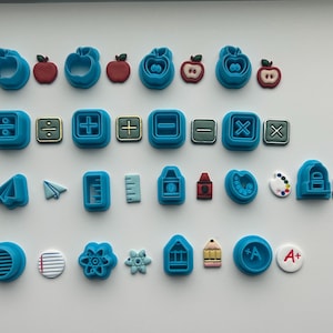 Op de afbeelding: Een verzameling blauwe plastic koekjesvormen en de bijbehorende afgewerkte vormen van klei of fondant. De set bevat schoolthema-ontwerpen zoals appels, wiskundige symbolen, papieren vliegtuigjes, linialen, kleurpotloden, verfpaletten en rugzakken.