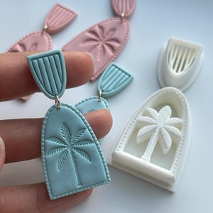 May include: Polymer clay earrings in pastel shades. The earrings feature a palm tree design and a textured finish. The earrings are in light blue and pink, with a white palm tree mold. The earrings are connected with silver-coloured jump rings.