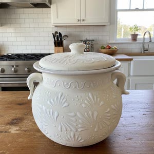 May include: A white ceramic lidded pot with embossed floral and wave patterns. The pot has two curved handles and a knob on the lid. The kitchenware is sitting on a wooden surface.