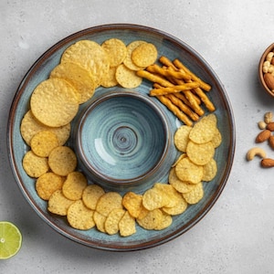 May include: A blue and brown ceramic serving platter filled with tortilla chips, pretzel sticks, and a central dip bowl. A small wooden bowl of mixed nuts and a sprig of rosemary are also present.