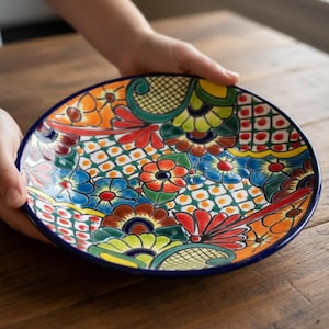 May include: A colorful ceramic plate with a vibrant floral design. The plate features a blue rim and a detailed pattern of flowers in shades of orange, red, blue, and green. The plate is held by two hands, showcasing the intricate artwork.