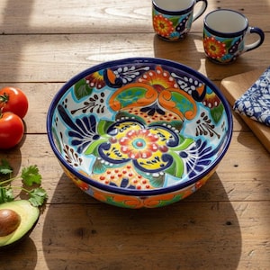 May include: A large, hand-painted ceramic bowl with a vibrant floral design in orange, blue, yellow, and green. The bowl is surrounded by fresh tomatoes, cilantro, and an avocado half. Two matching mugs and a patterned napkin are also present.