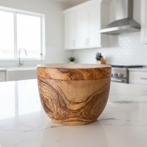 May include: A wooden bowl with a lid, showcasing a natural wood grain pattern. The bowl is light brown with darker brown swirls, and the lid is a lighter shade of brown. The bowl is sitting on a white countertop in a kitchen setting.