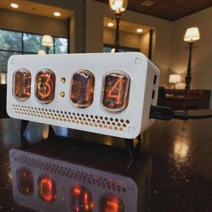 Könnte beinhalten: Eine weiße Digitaluhr mit leuchtend orangefarbenen Ziffern, die "03:14" anzeigen. Die rechteckige Uhr hat einen perforierten unteren Rand und wird von schwarzen Beinen getragen. Ein schwarzes USB-Kabel ist an der Seite angeschlossen. Ca. 15 cm lang.