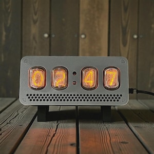 Könnte beinhalten: Eine graue Digitaluhr, die "00:00" anzeigt, mit vier leuchtenden orangefarbenen Röhren. Die Uhr hat eine rechteckige Form mit einem perforierten unteren Bereich und steht auf einem schwarzen Ständer. Der Hintergrund ist eine Holzwand und ein Holzboden.