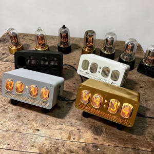 Könnte beinhalten: Eine Sammlung digitaler Uhren im Retro-Stil mit leuchtend orangefarbenen Ziffern. Die Uhren haben rechteckige Gehäuse in verschiedenen Farben, darunter Schwarz, Weiß, Gold und Grau. Mehrere Nixie-Röhren sind ebenfalls zu sehen.