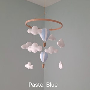 May include: A baby mobile with a wooden ring, white felt clouds, and two pastel blue hot air balloons with woven baskets. The text "Pastel Blue" is displayed below the mobile.