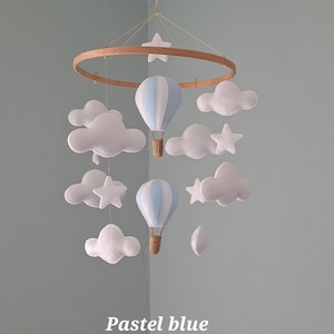 May include: A hanging mobile featuring two pastel blue and white striped hot air balloons with brown woven baskets, suspended from a circular wooden hoop. The mobile includes several white felt clouds and stars hanging at varying heights.