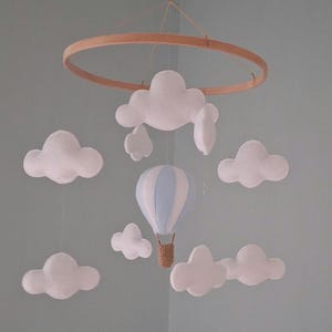 May include: A nursery mobile with a wooden hoop, white felt clouds, and a blue and white hot air balloon. The mobile is suspended by a string. The design is suitable for a baby's room or a child's bedroom.