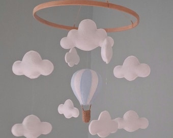 Puede incluir: Móvil para bebé con un aro de madera, nubes blancas y un globo aerostático azul y blanco. El móvil está suspendido por una cuerda. Ideal para la habitación del bebé.