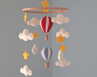 Puede incluir: Un móvil colgante con un marco circular de madera y dos globos aerostáticos, uno rojo y blanco y otro azul y blanco, suspendidos por hilos. El móvil incluye varias nubes de fieltro blanco y estrellas de fieltro amarillo a varios niveles.