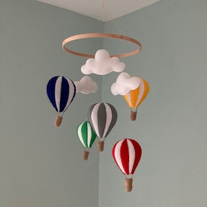 Può includere: Un mobile a cerchio di legno bianco con nuvole e mongolfiere in feltro di vari colori, tra cui rosso, blu, verde, giallo e grigio.