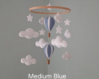 Könnte beinhalten: Ein Baby-Mobile mit einem Holzring, weißen Wolken, blau-weiß gestreiften Heißluftballons und weißen Sternen. Das Mobile ist mit "Medium Blue" beschriftet.