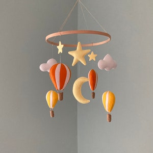 May include: A hanging mobile featuring a circular wooden frame with suspended felt elements including orange and white hot air balloons, yellow and white hot air balloons, a large yellow star, two small yellow stars, a yellow crescent moon, and two light grey clouds.