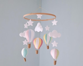 May include: A baby mobile featuring a wooden ring with felt stars and clouds. Five colorful hot air balloons in pastel shades of pink, blue, yellow, and green hang below. The balloons have woven baskets. The mobile hangs against a light blue wall.