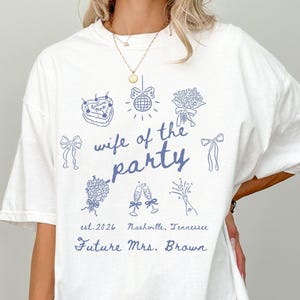 Puede incluir: Camiseta blanca con diseño azul que presenta el texto "wife of the party" y varias ilustraciones. Incluye el texto "est. 2026 Nashville, Tennessee Future Mrs. Brown". Una camiseta informal y de gran tamaño.