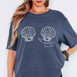 以下が含まれることがあります： 白い貝殻のデザインが施されたデニムブルーのTシャツ。シャツには「Olivia's Bachelorette Miami, Florida」の文字が入っています。このシャツは半袖のカジュアルスタイルです。