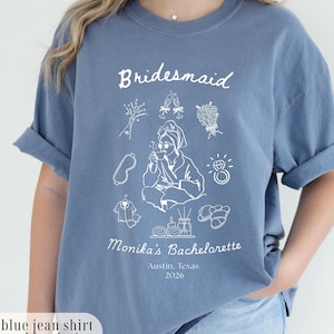 May include: A blue denim-style t-shirt with a white graphic. The shirt displays the word "Bridesmaid" and various illustrations, including a bride, flowers, and a ring. Below, it reads "Monika's Bachelorette, Austin, Texas, 2026."
