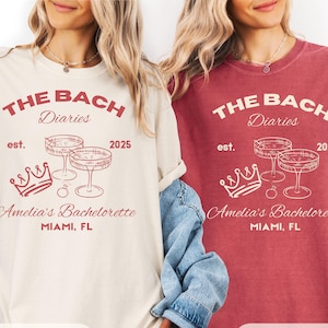 May include: Two t-shirts, one ivory and one crimson, display the text "THE BACH Diaries" with "est. 2025" and "Amelia's Bachelorette MIAMI, FL." The design includes champagne glasses and a crown. The ivory shirt has a crimson design, and the crimson shirt has an ivory design.
