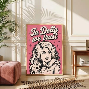 May include: A rectangular wall art print featuring a stylised black and white illustration of a woman with voluminous wavy hair and hoop earrings. The background is pink with the text "In Dolly we trust" in a bold, dark script font above the portrait.