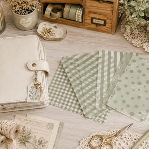 May include: A cream-colored leather planner with a gold snap closure and a floral charm, accompanied by several translucent sage green patterned binder inserts featuring stripes, gingham, and polka dots, alongside stationery supplies.