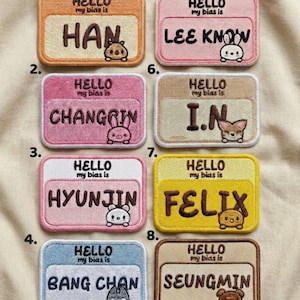 Puede incluir: Una colección de ocho parches bordados con nombres en varios colores, cada uno con un nombre y un pequeño personaje de dibujos animados. Los parches tienen forma rectangular con esquinas redondeadas y el texto "HELLO my bias is" encima del nombre.