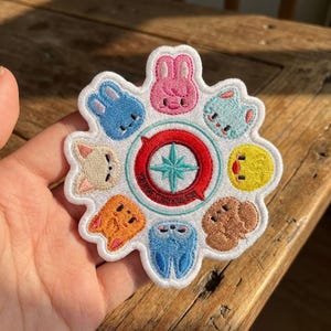 May include: Embroidered patch featuring a circular design with cartoon animal faces in various colors, including blue, pink, and yellow. The center has a compass rose design with text that reads "YOU MAKE STRAY KIDS STAY".