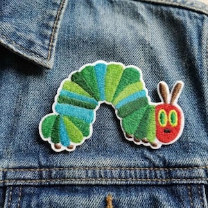 May include: Embroidered patch featuring a colorful caterpillar design with green, blue, and red accents. The patch is attached to a denim jacket, showcasing its vibrant colors and detailed stitching. A fun, whimsical accessory.