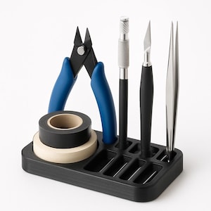 May include: A black organizer holds various craft tools, including blue-handled pliers, a silver-handled craft knife, a black-handled blade, and tweezers. Rolls of black and tan tape are also present. The tools are arranged on a black, rectangular base.